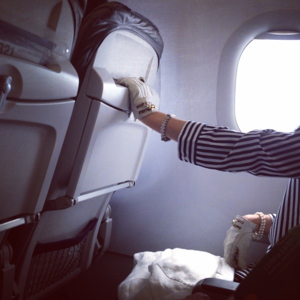 Aircraft cabin interior: a hand in a lace fingerless glove with rings and a pearl bracelet resting on the seat back in front, the oval window burnt out white