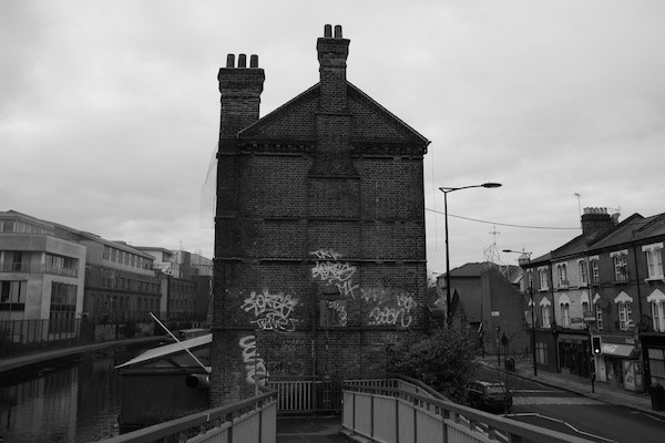 Black-and-white photograph of a tall brick end-of-terrace building with two chimney stacks and graffiti on its lower wall, standing beside a canal
