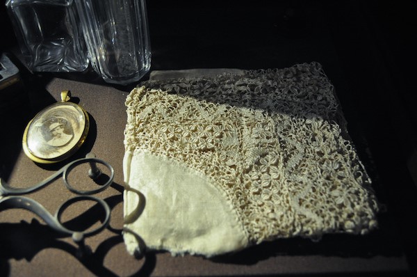 Still life in a shaft of light: a piece of antique cream lace on brown paper beside ornate metal scissors, a small oval locket and two cut-glass tumblers