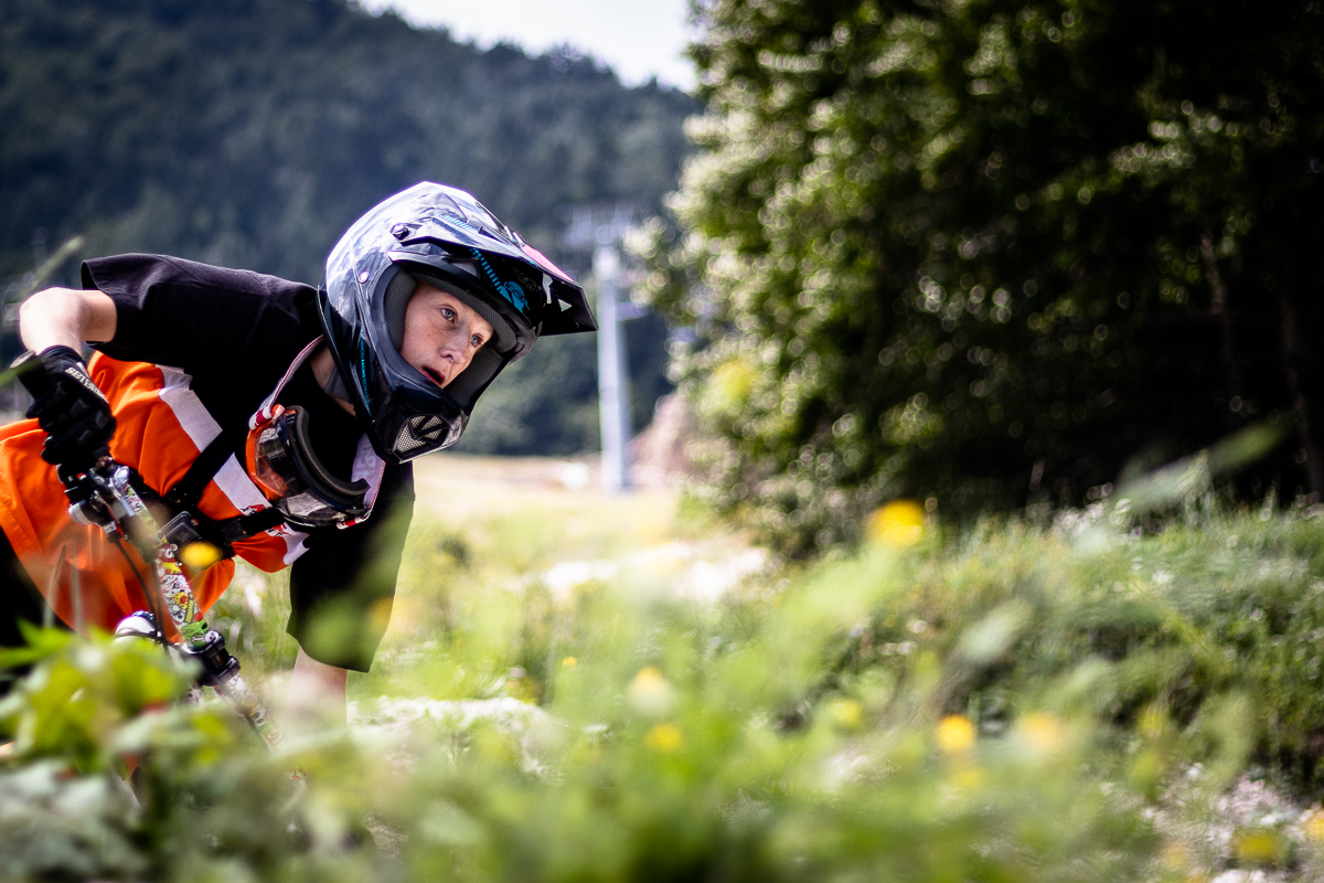Young downhill rider in an orange jersey crouched low over the handlebars at Bike Park Kranjska Gora, with a chairlift pylon behind