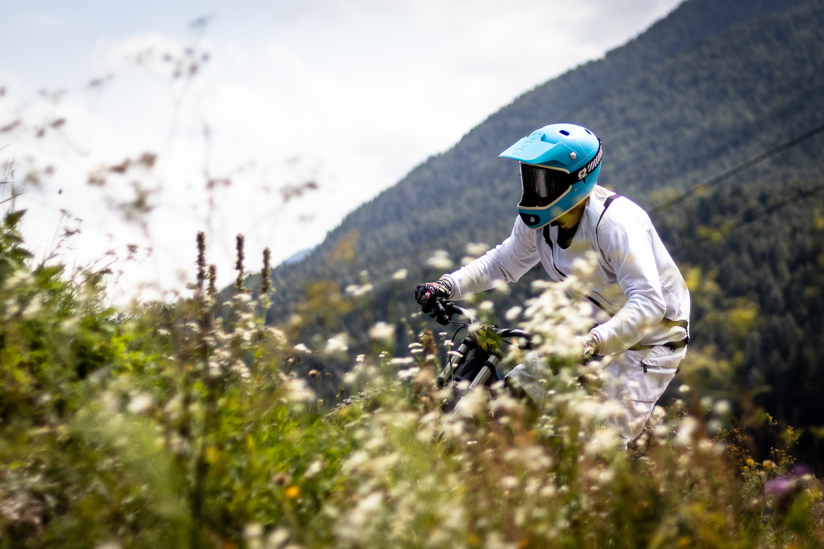 Downhill mountain biker in a white jersey and light blue full-face helmet riding through white wildflowers at Bike Park Kranjska Gora