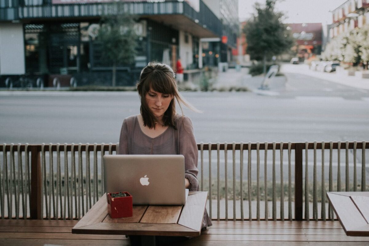 A woman working on a laptop at an outdoor cafe table beside a quiet street, the screen turned away from the camera