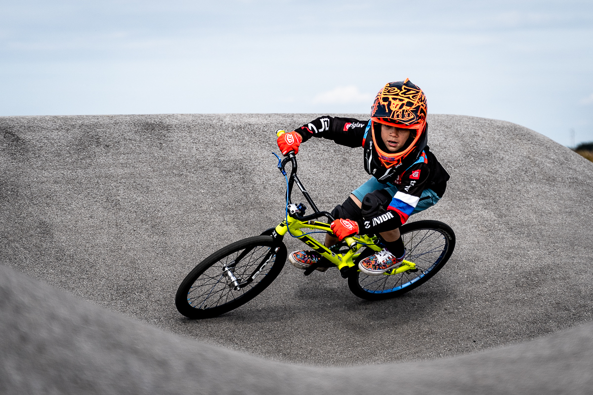 Young BMX rider in an orange helmet leaning through a grey asphalt berm at Pump Track Odranci