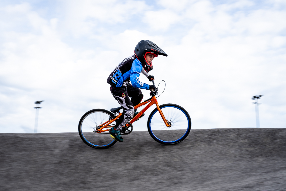 BMX racer wearing number 12 crossing a roller at Pump Track Odranci, with the asphalt streaked by motion blur