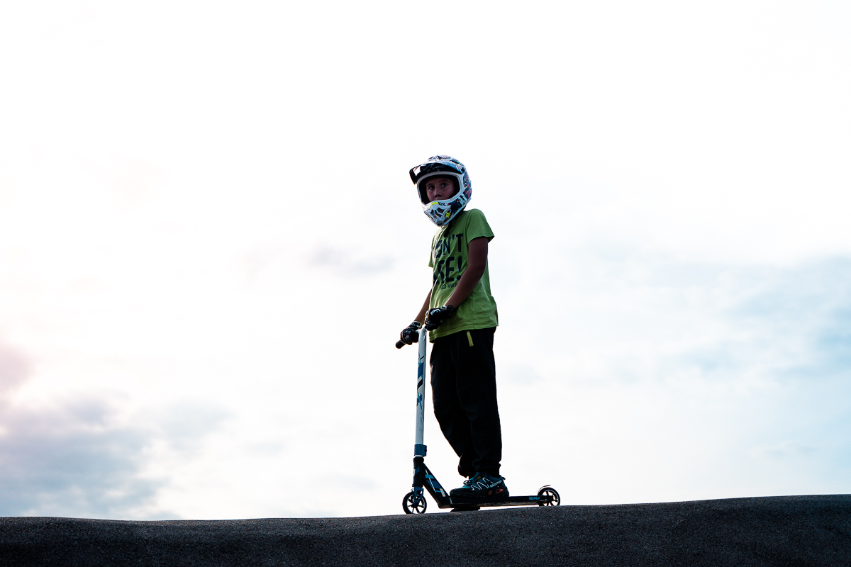 Child in a green shirt and white full-face helmet standing on a scooter on a crest at Pump Track Odranci