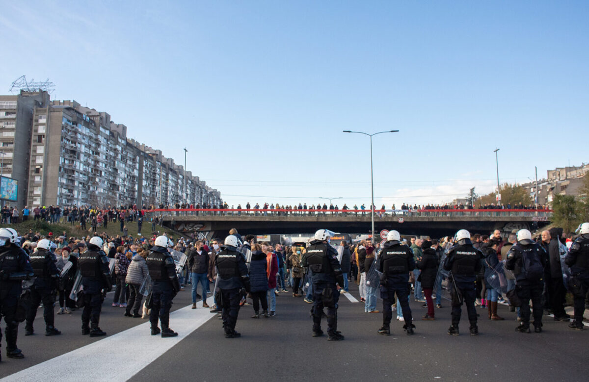 A line of riot police in helmets blocking a Belgrade highway, with protesters gathered on the bridge behind them under a clear blue sky