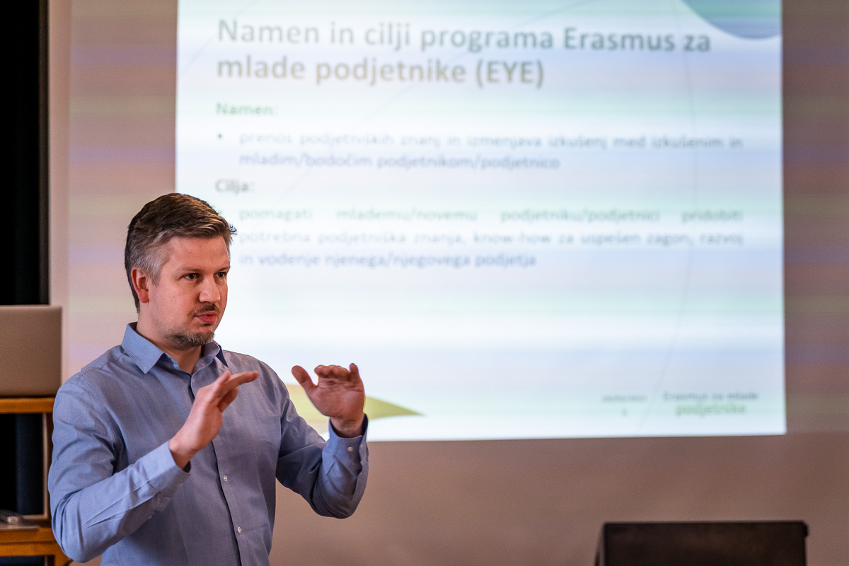 Speaker in a blue shirt presenting a slide titled Namen in cilji programa Erasmus za mlade podjetnike at MIKK Murska Sobota