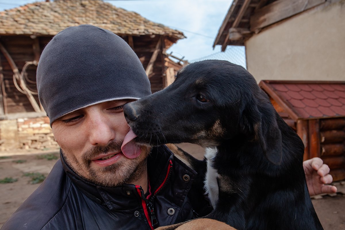 A man in a beanie laughing as a black and tan dog licks his face, rustic farm buildings behind them