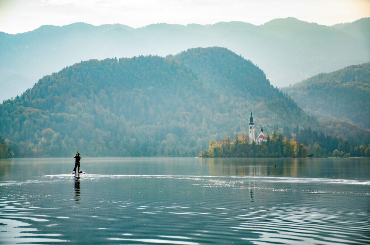 A lone paddleboarder crossing the still surface of Lake Bled at dawn, with the island church and layered misty hills behind