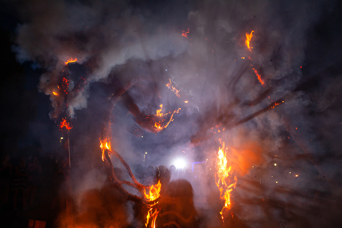 A large wicker or straw effigy burning at night, flames and sparks lighting the smoke as silhouetted figures watch
