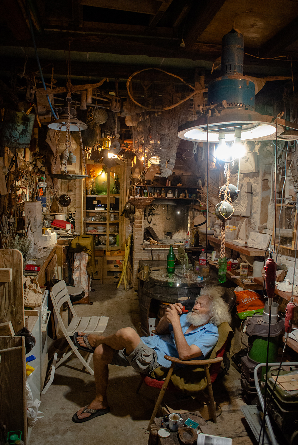 An elderly sculptor with wild white hair and beard smoking a pipe in a cluttered workshop crowded with tools, nets and old bottles, lit by a single overhead lamp