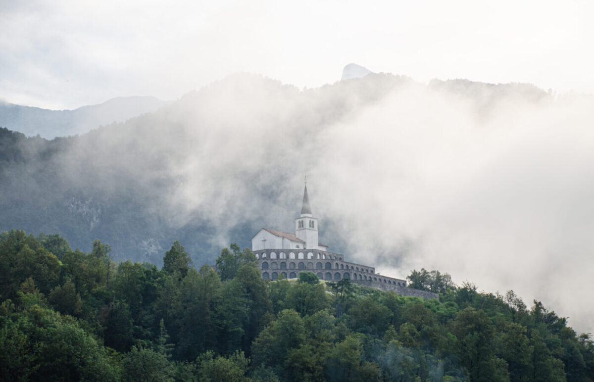 The Italian Charnel House above Kobarid: a white church with a spire on tiered arcaded terraces, half wrapped in low cloud on a wooded hill