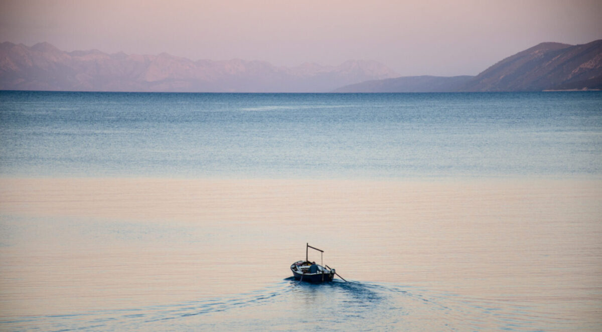 A small wooden boat leaving a wake across a calm pastel sea at dawn, with layered mountain ridges on the horizon