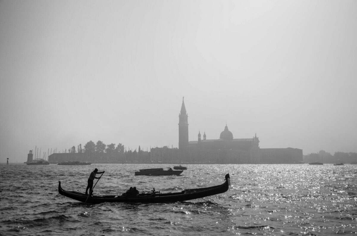 Black-and-white photograph of a gondolier poling across the Venetian lagoon, with the campanile and dome of San Giorgio Maggiore in the haze behind