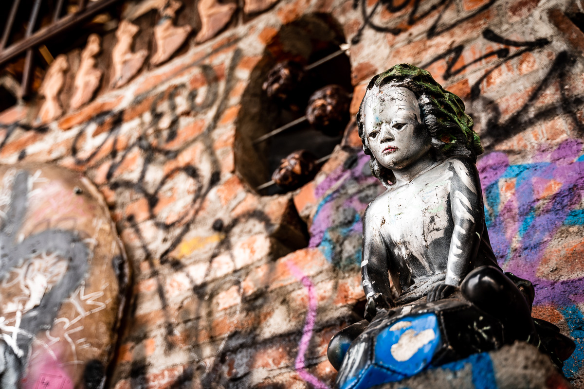 Weathered sculpture of a small girl against a brick and graffiti wall at Metelkova