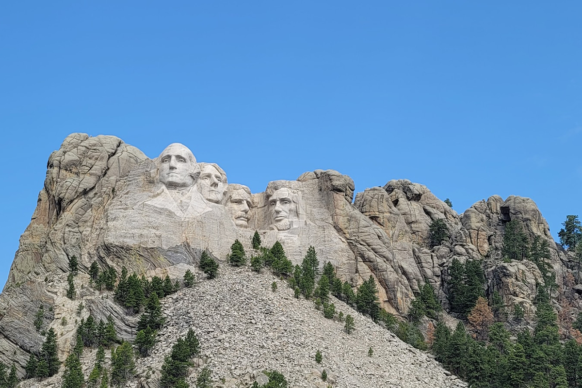 Wide view of Mount Rushmore above a broad talus slope and pine forest