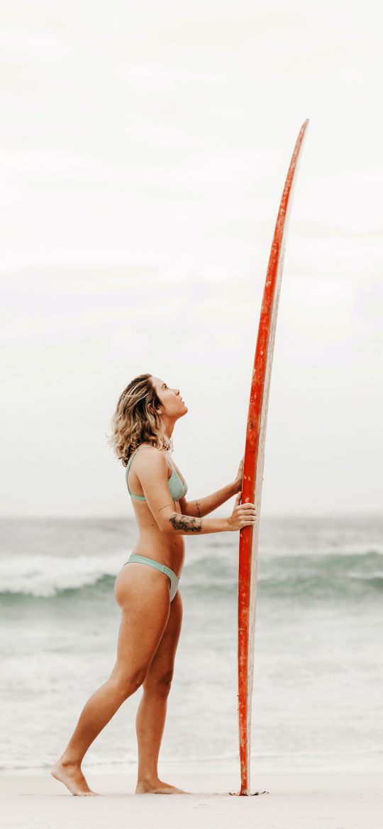 Woman standing beside the sea with a tall red surfboard