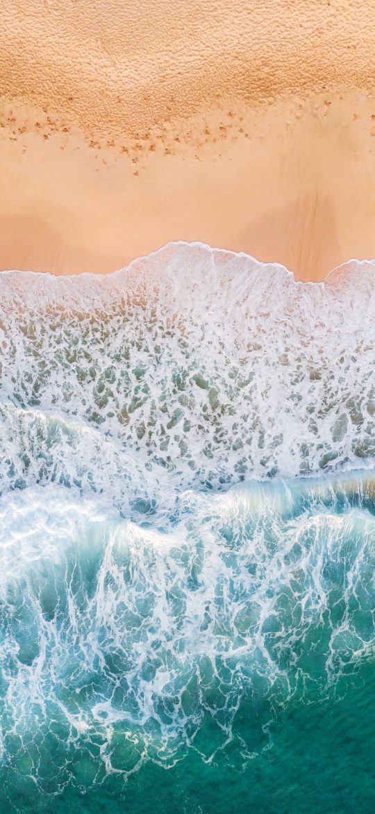 Vertical aerial wallpaper of a wave washing across pale sand