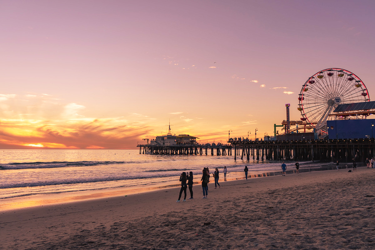 People walking on Santa Monica Beach as the pier and Pacific Wheel extend into the ocean at sunset