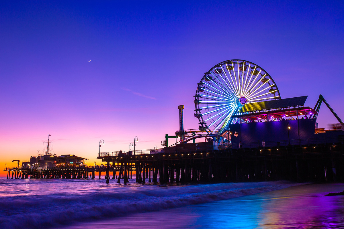 Santa Monica Pier during blue hour with the illuminated Pacific Wheel, a crescent moon and reflected colored light on the water