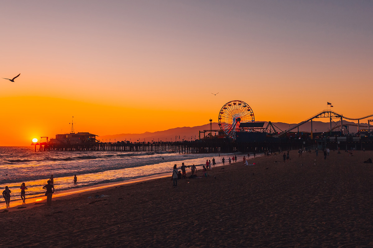 The setting sun beside Santa Monica Pier, with the Pacific Wheel and beachgoers in silhouette