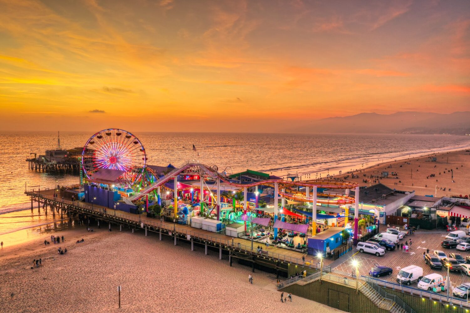 Pacific Park and Santa Monica Pier illuminated beneath an orange sunset, viewed from above