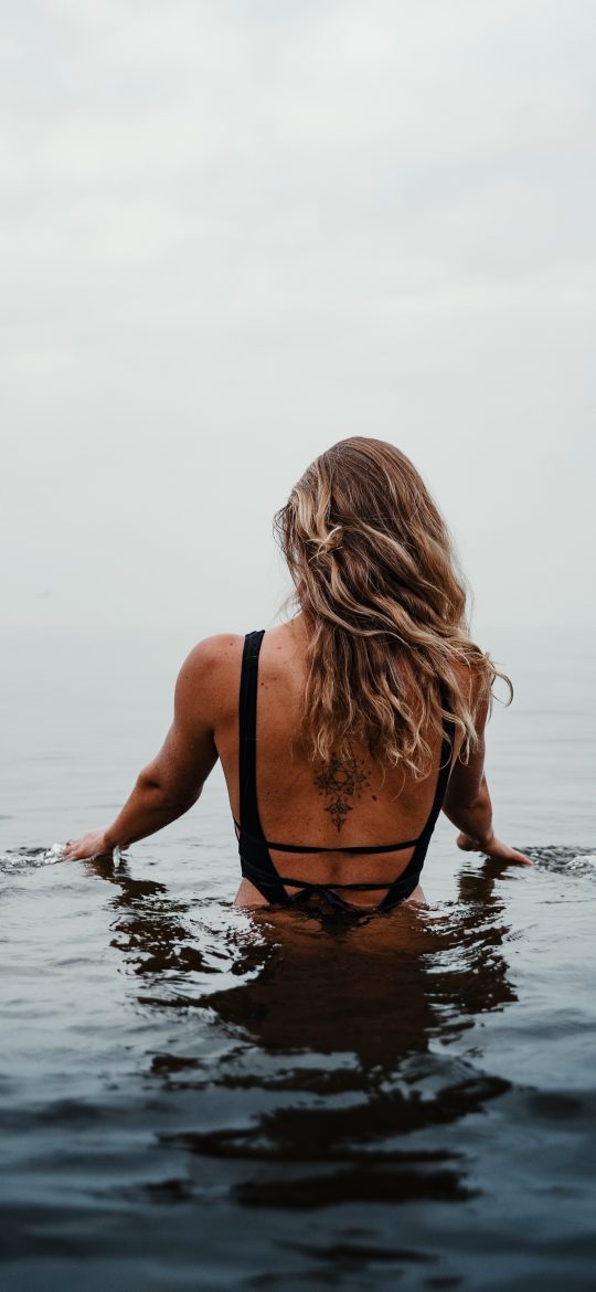 Woman seen from behind while standing in dark sea water beneath an overcast sky