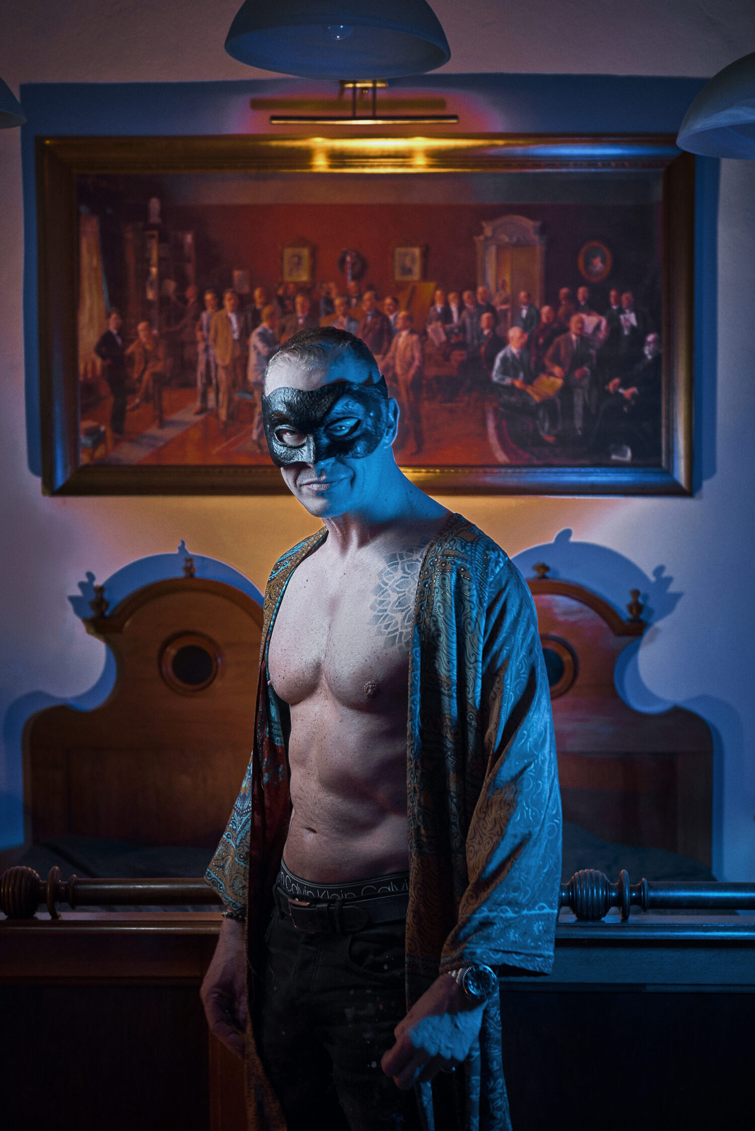 A man in a black Venetian masquerade mask and an open patterned silk robe, lit in blue, standing before a large framed group painting