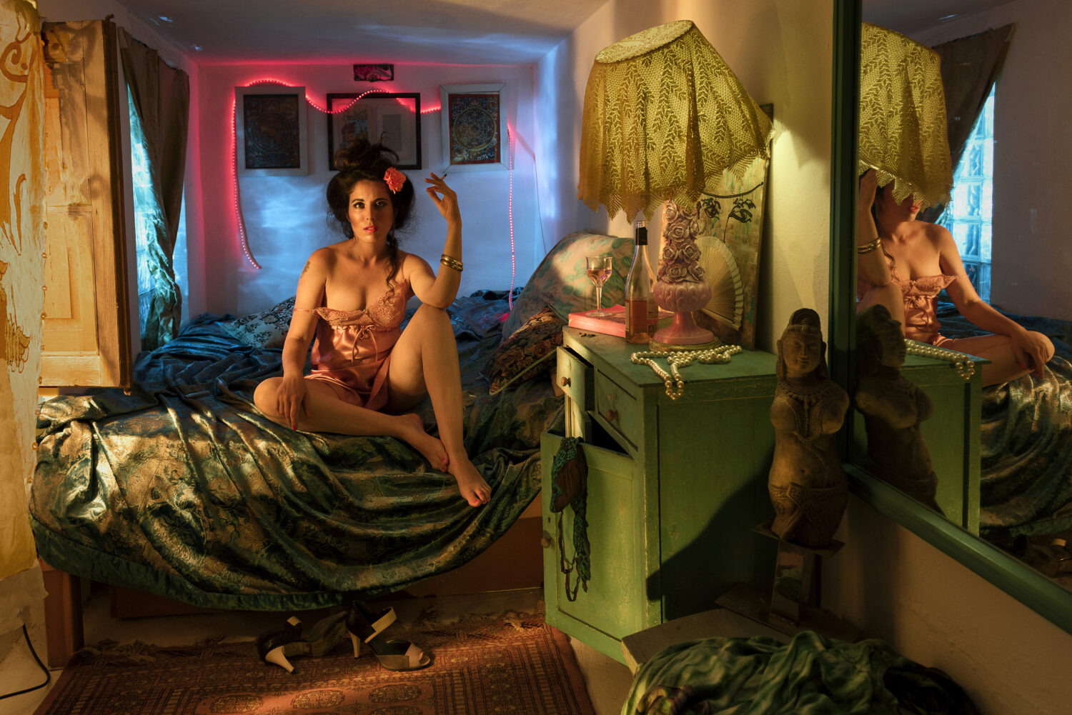 Wide bedroom scene: a woman in a peach silk slip sitting cross-legged on a green brocade bed, pink neon rope light on the wall behind, a lace-shaded lamp and tall mirror to the right