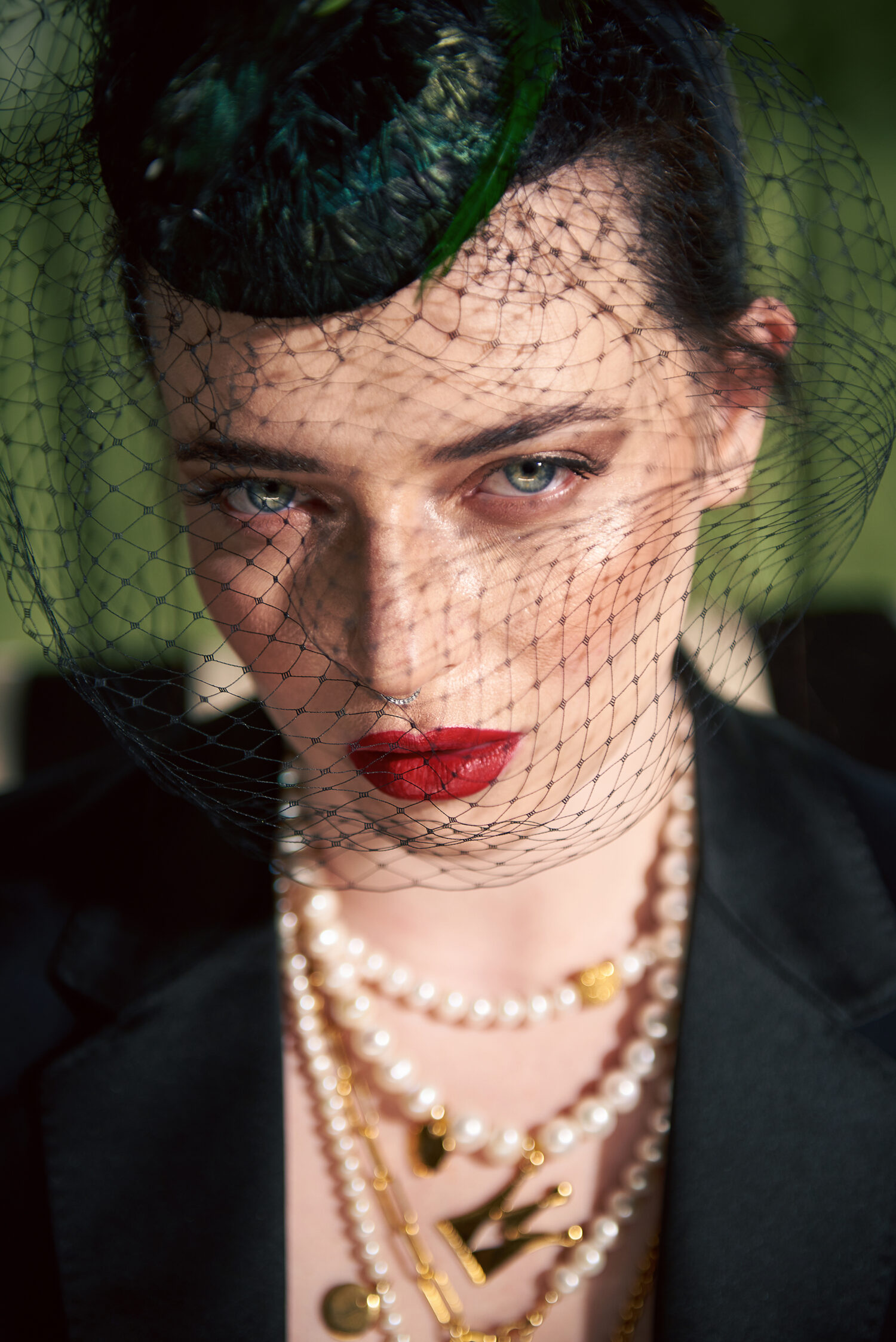 Close portrait of a woman in a black feathered fascinator with a birdcage veil across her face, red lipstick and layered pearl necklaces, against a green background